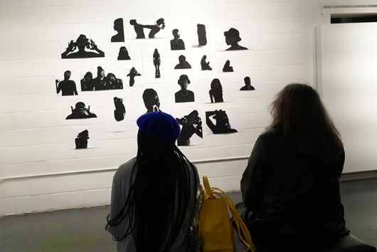 Two guests in the gallery, sitting on a bench while pondering the artwork of silhouetted bodies on the wall.
