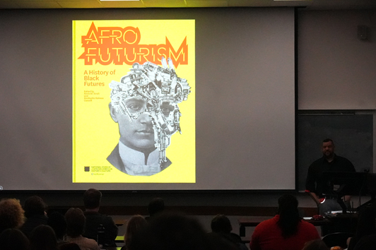 Artist Wayne Hodges giving a lecture in front of a classroom. On screen is an image of a book cover called Afrofuturism: A History of Black Futures.