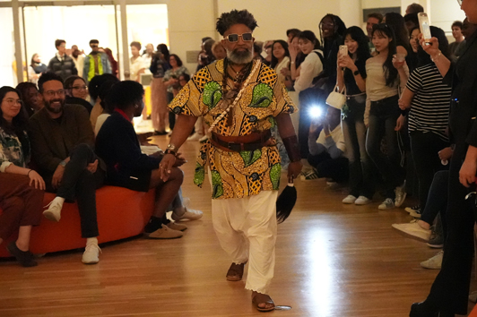 A fashion show model wearing afrofuturist regalia strutting in front of a crowd of onlookers taking photos.