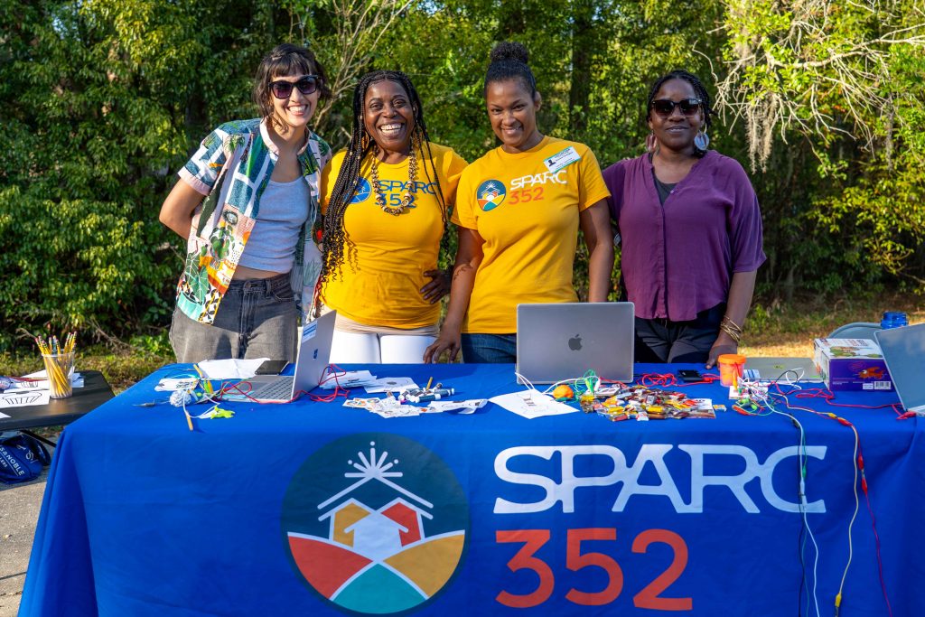 Four artist facilitators smile at the camera behind a blue tablecloth that has the sparc logo on it.