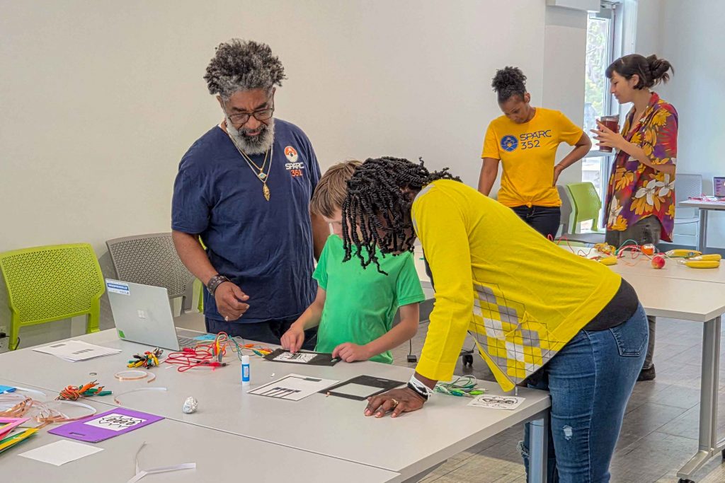 Two artist facilitators instructing a young boy on a project using paper, wires and a laptop.
