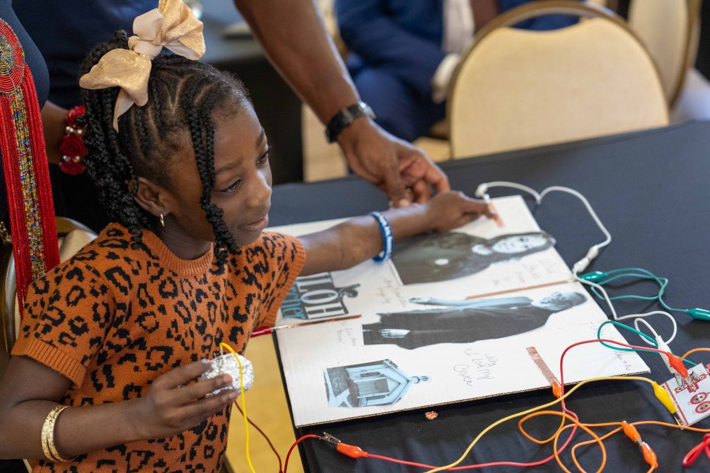 A young girl touches an interactive collage featuring local leaders. The collage has copper tape and wires connecting it to a makey makey