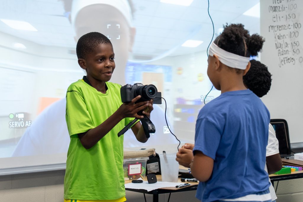 A young boy holds a camera pointed at another student.