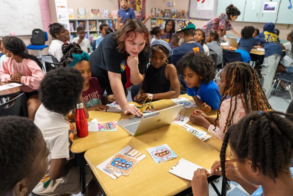 A facilitator at a group of desks and students, the students lean over to look at a laptop screen. There are papers with colorful creatures and copper tape, one is connected with wires to the laptop.