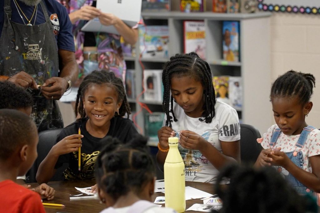 Two young girls are looking down in concentration on the project they are working on while a third girl smiles at someone across the table.